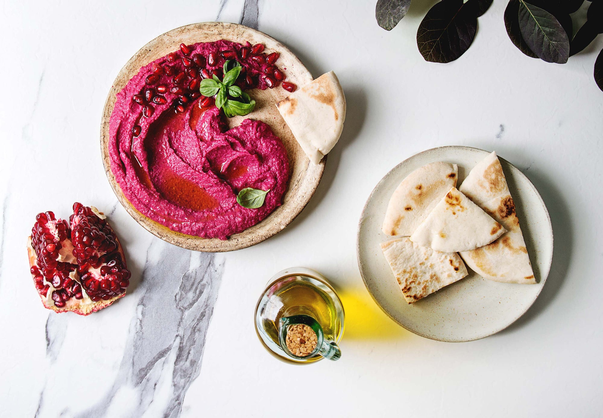 A marble countertop with a plate of beet hummus, a plate of pita bread slices, a pomegranate, and a cruet of olive oil.