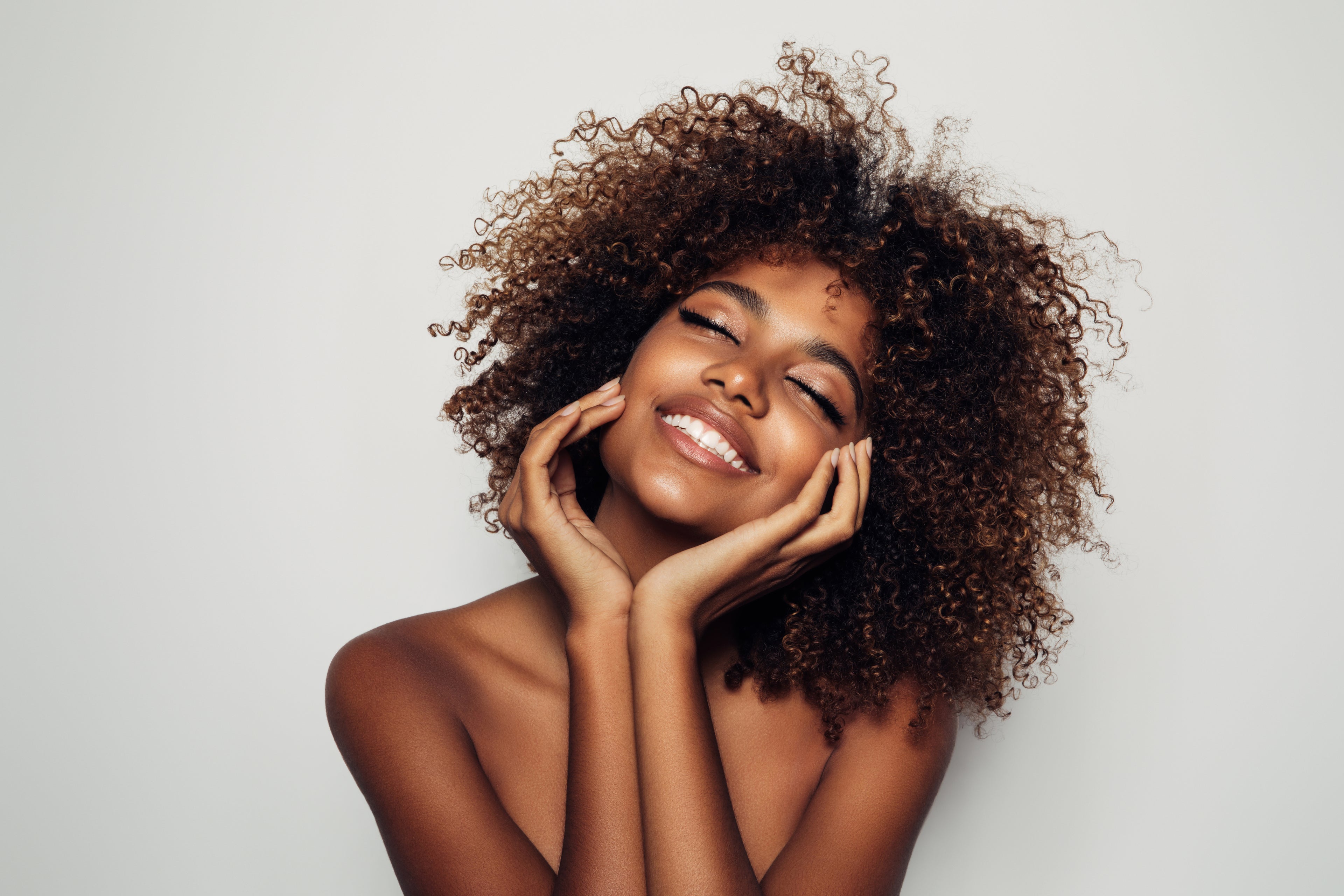 A woman with clean, glowing skin touching her face, smiling and tilting her head upwards.