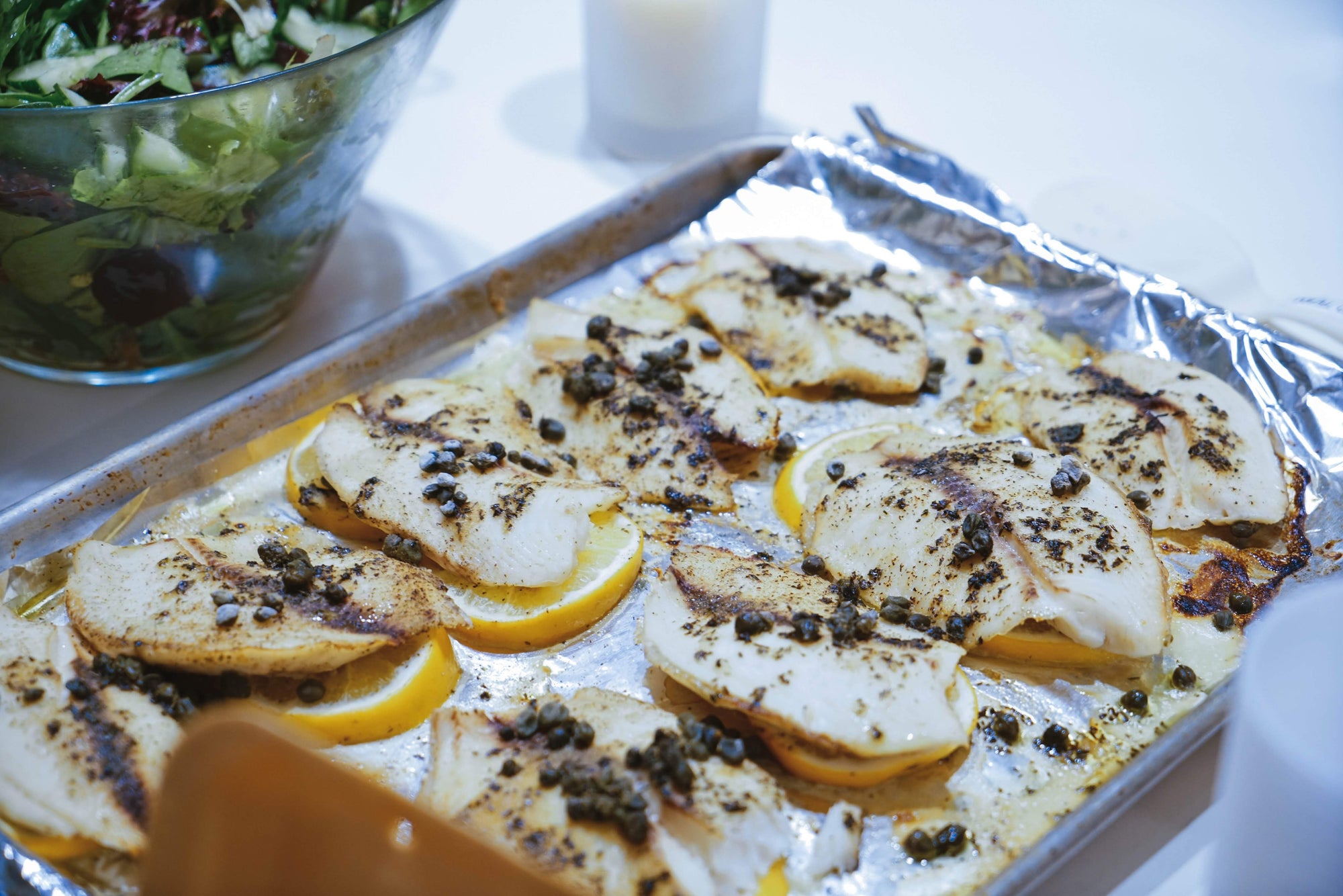 A sheet pan topped with aluminum foil, tilapia filets, capers, and lemon slices.