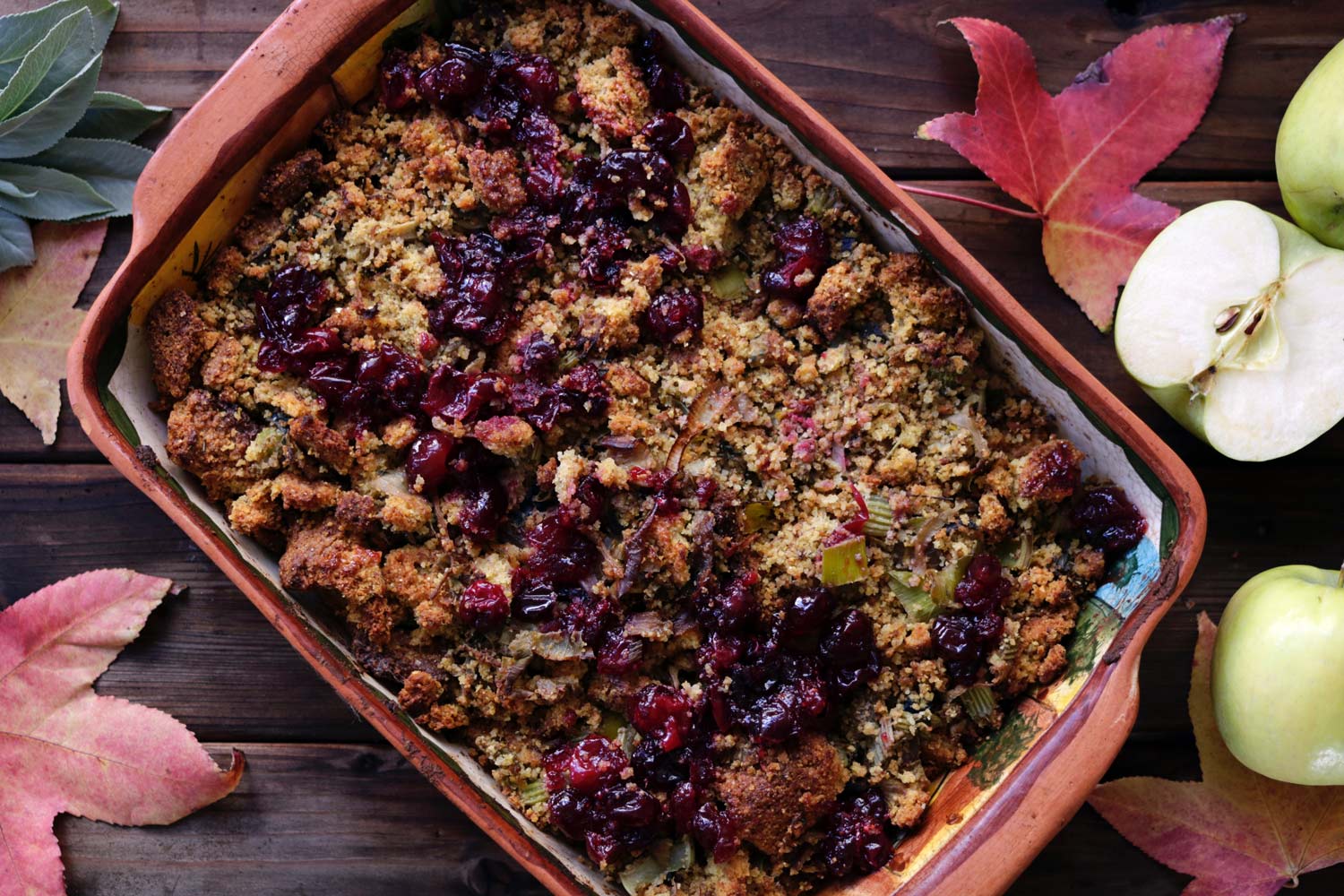 A baking dish of Thanksgiving stuffing on a wooden table surrounded by green apples and maple leaves.