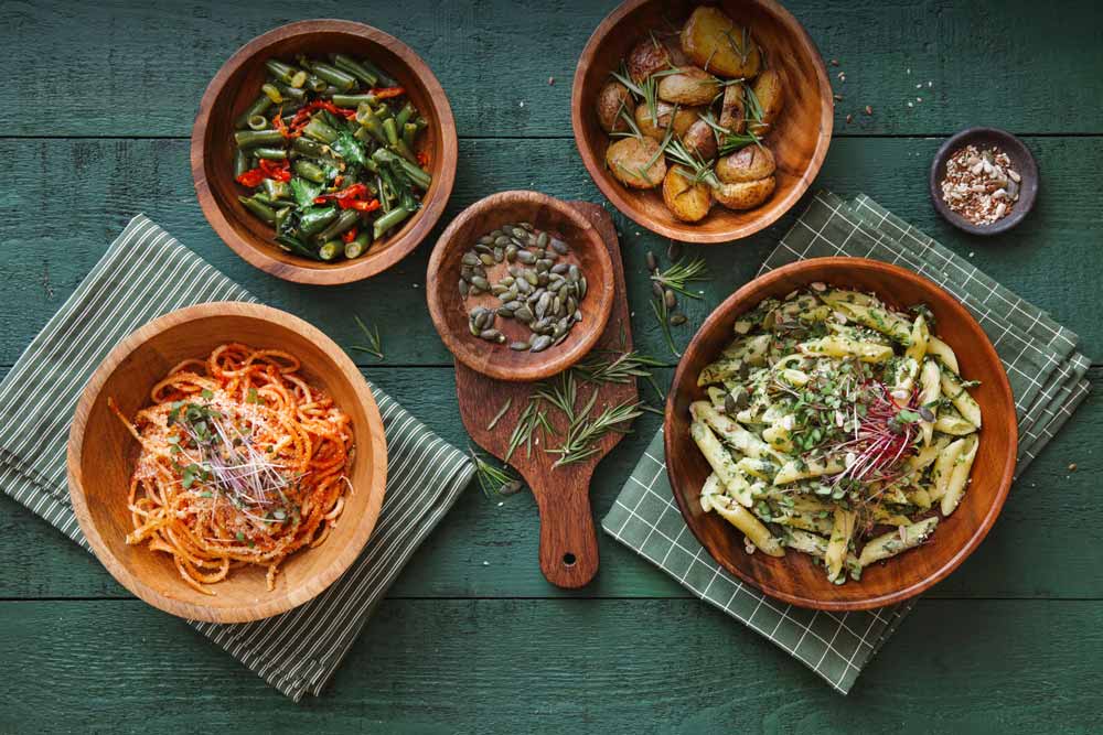 Top view image of a wooden table stained green, with 5 bowls of food: pasta, vegetables, and potatoes.